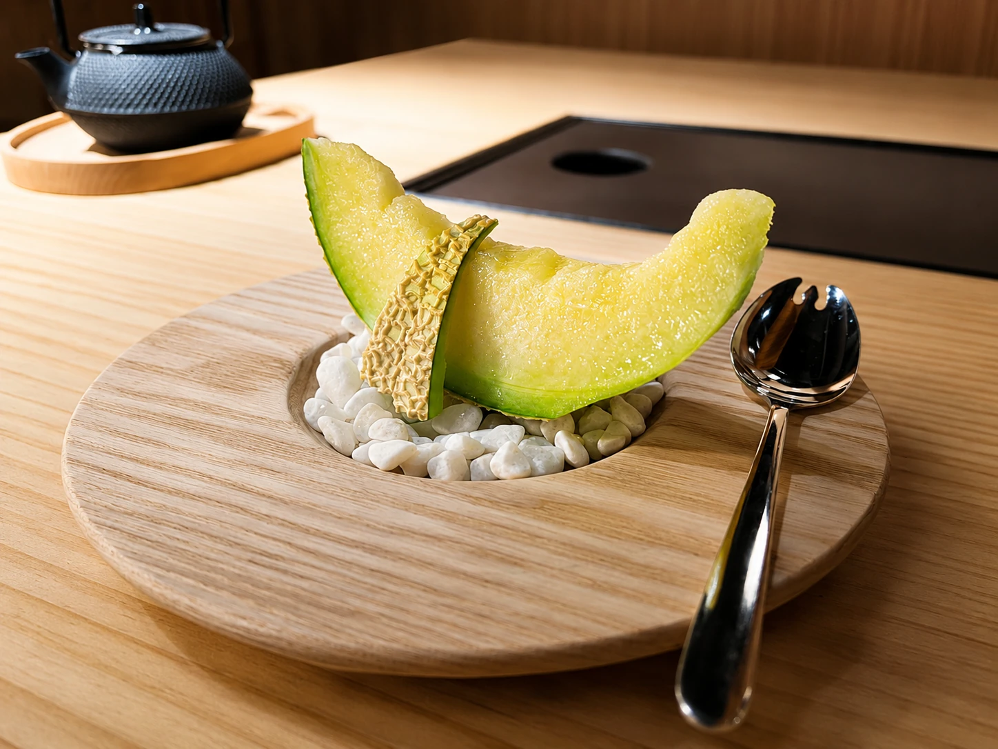A slice of Crown Melon plated on white pebbles in a carved wooden dish, with a cast-iron teapot on the counter behind it